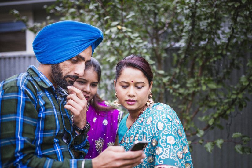 Sikh man looking at his phone thoughtfully, next to two women dressed in sari.