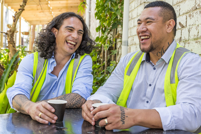 Two male colleagues in high-vis vests having a coffee and laughing at an outdoor café.
