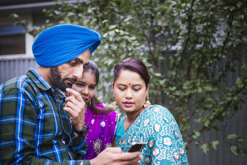 A man looking at his phone thoughtfully, next to two women dressed in sari.
